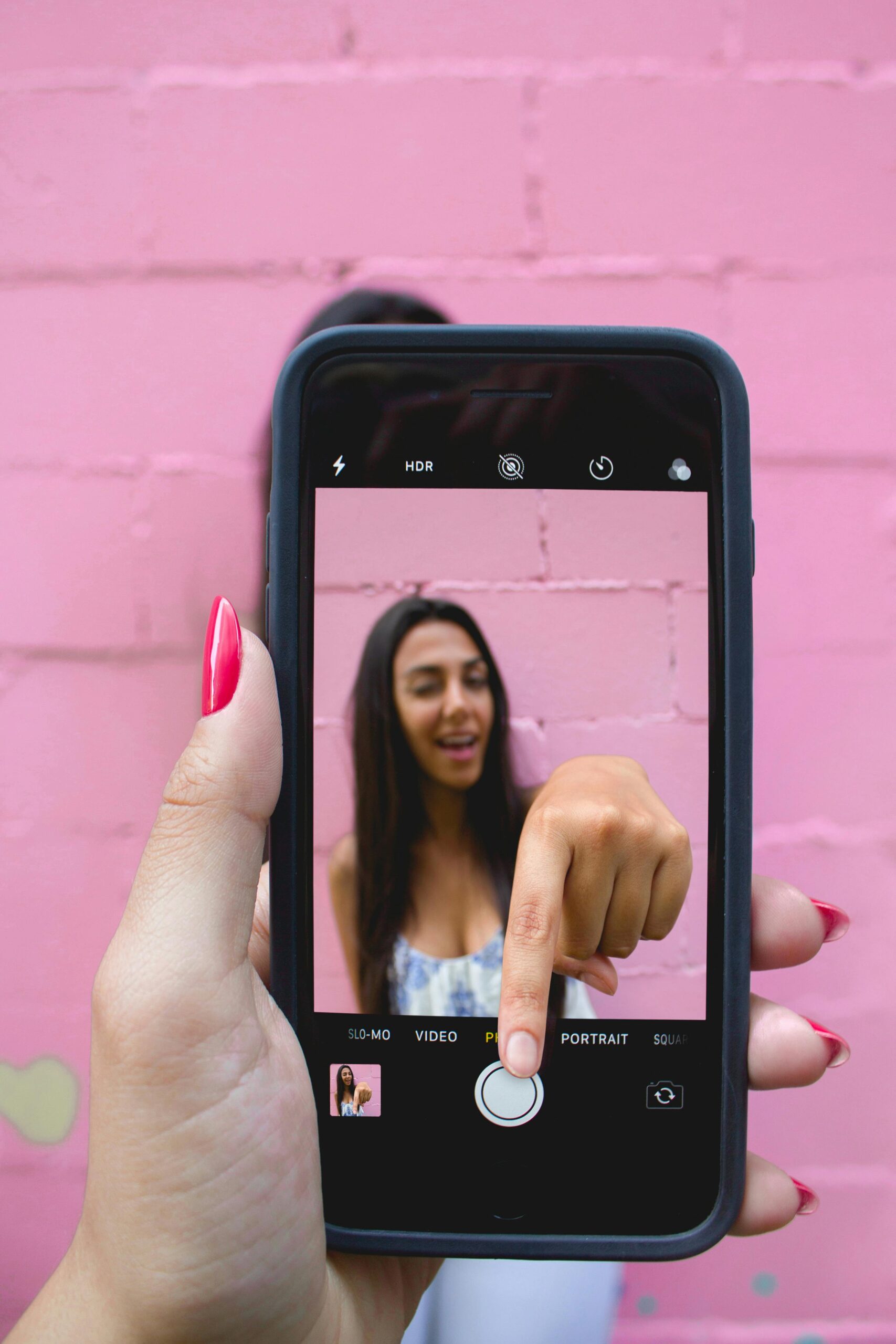 A woman captures her selfie using a smartphone against a vibrant pink wall.